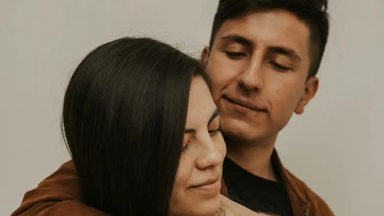A couple stands close together, with one person embracing the other from behind.