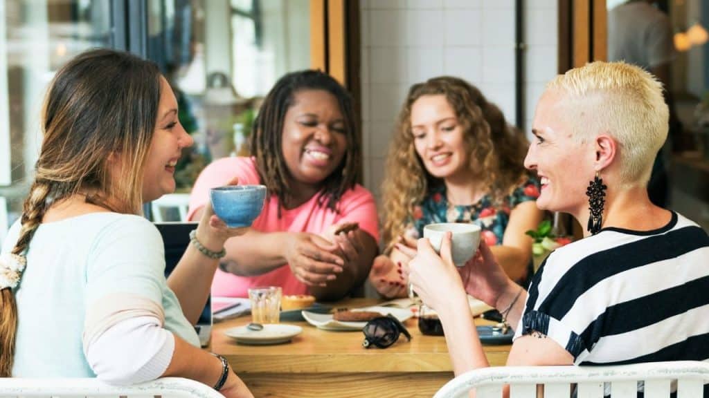 Four women sit around a wooden table, laughing and holding small blue and white bowls.