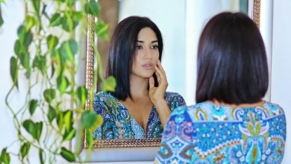A woman in a blue patterned dress looks at her reflection in a framed mirror.