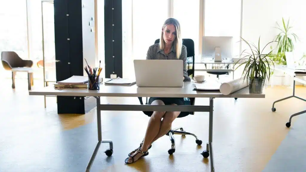 A woman sits at a large desk in a bright, modern office using a laptop.