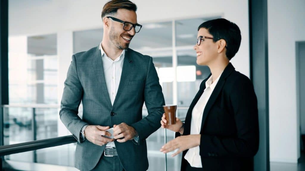 A man and woman in business attire smile and talk while holding a coffee cup.