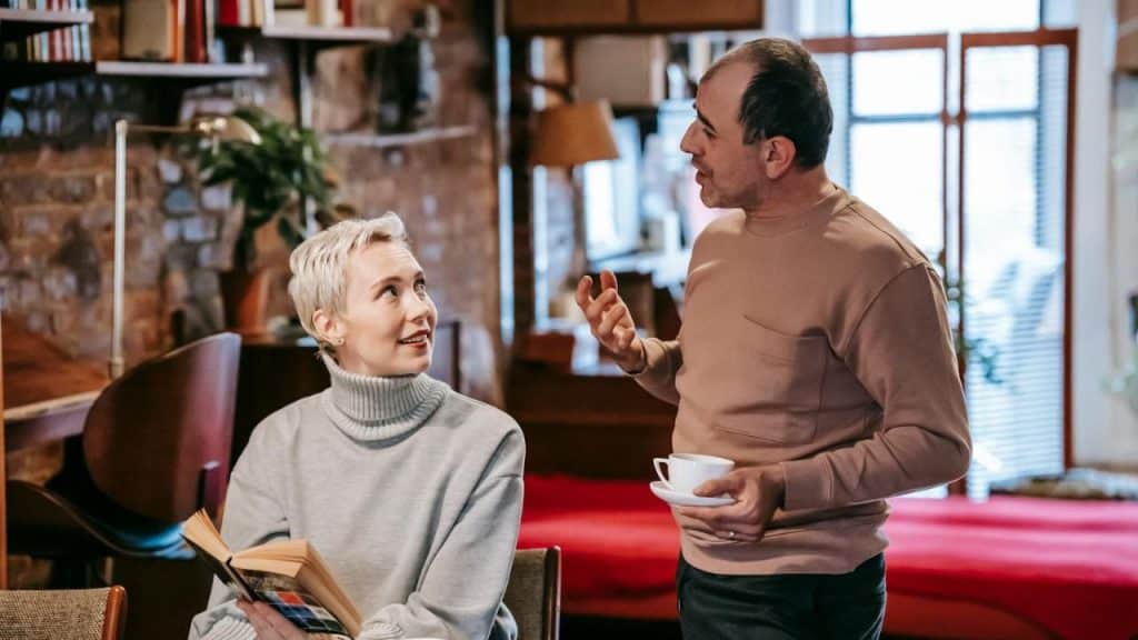 A woman with short blonde hair holds a book and looks at a gesturing man.