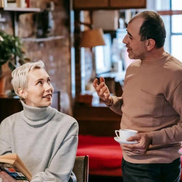 A woman with short blonde hair holds a book and looks at a gesturing man.