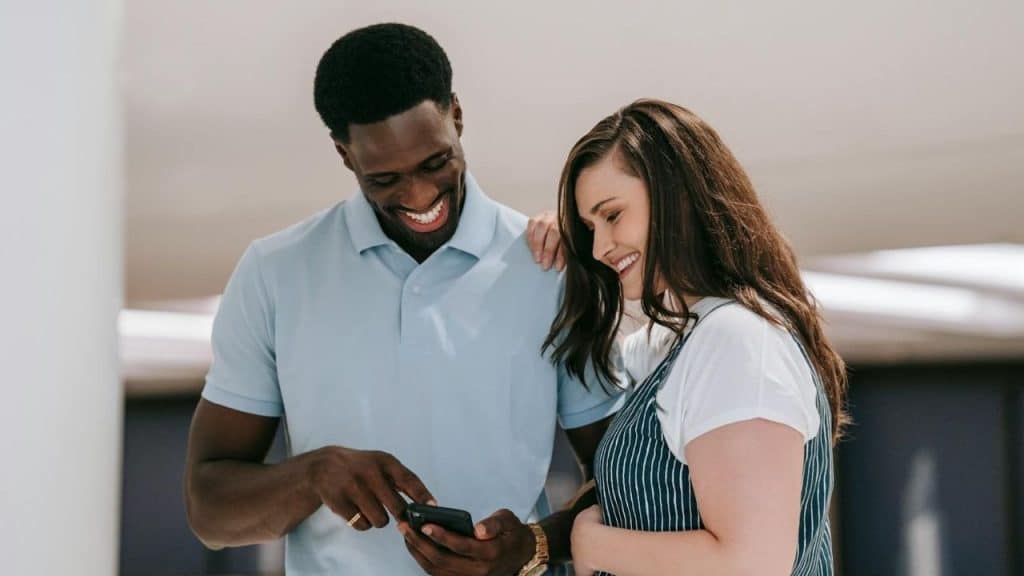 A man and woman stand close together while smiling and looking down at a smartphone.