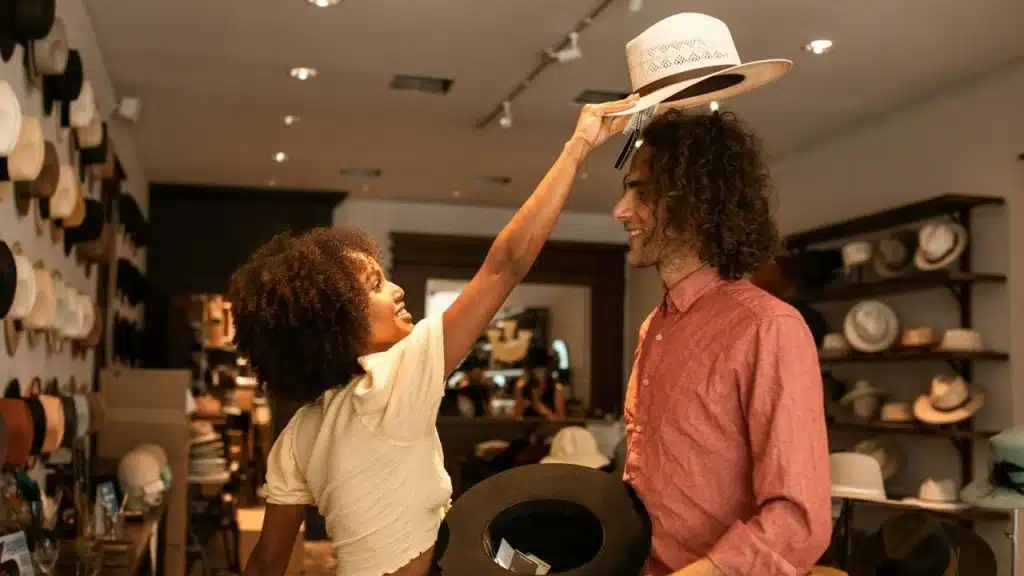 A woman places a white hat on a man's head inside a shop filled with hats.
