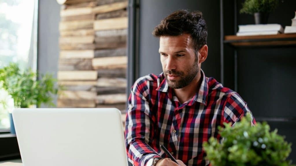 A man in a plaid shirt looks down at a laptop screen while holding a pen.