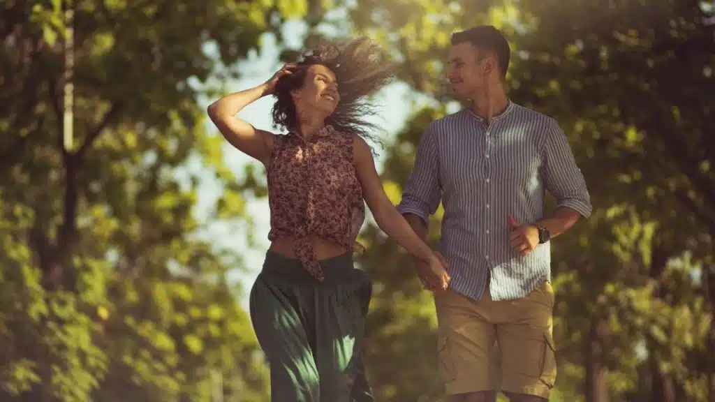 A man and woman walk hand-in-hand through a sunny park with green trees.