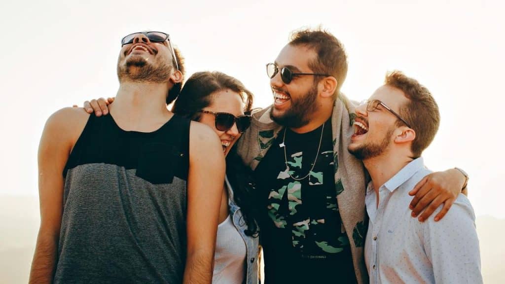 Four young adults with sunglasses laugh together outdoors with their arms around each other.