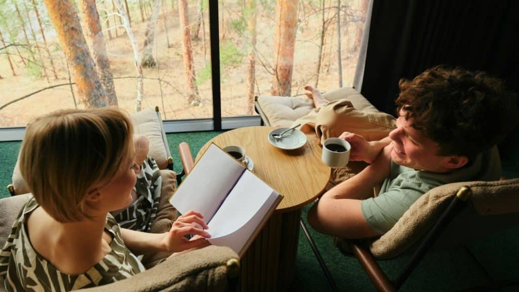 Two people sit in lounge chairs by a window, reading and drinking coffee.