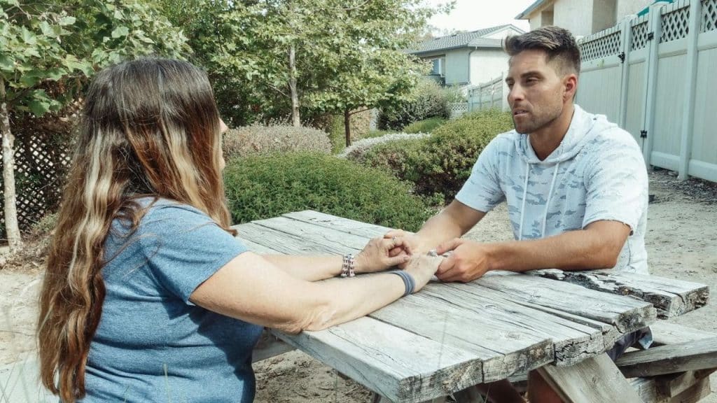 A man and woman sit at a wooden picnic table outdoors holding each other's hands.