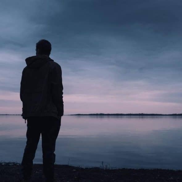A silhouetted person standing by a calm lake under a cloudy dusk sky.