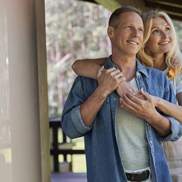 A smiling couple standing on a balcony, where the woman has her arms around the man’s shoulders, who holds them.