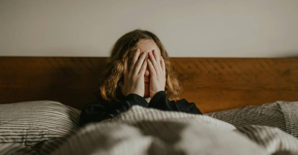 A person sitting in bed covering their face with both hands.