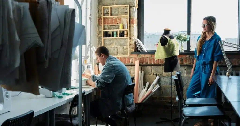 A fashion studio where a person sews at a desk while another stands nearby.