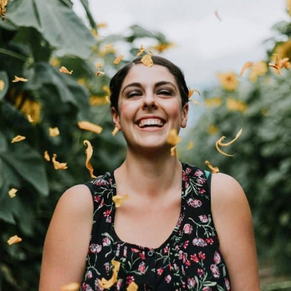 A woman smiling while standing between trees as withered leaves fall on her.