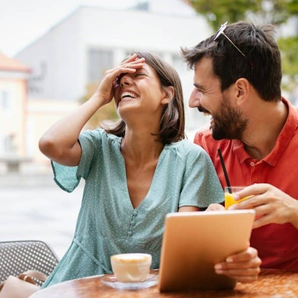 A couple sitting at a table and laughing while looking at a tablet.