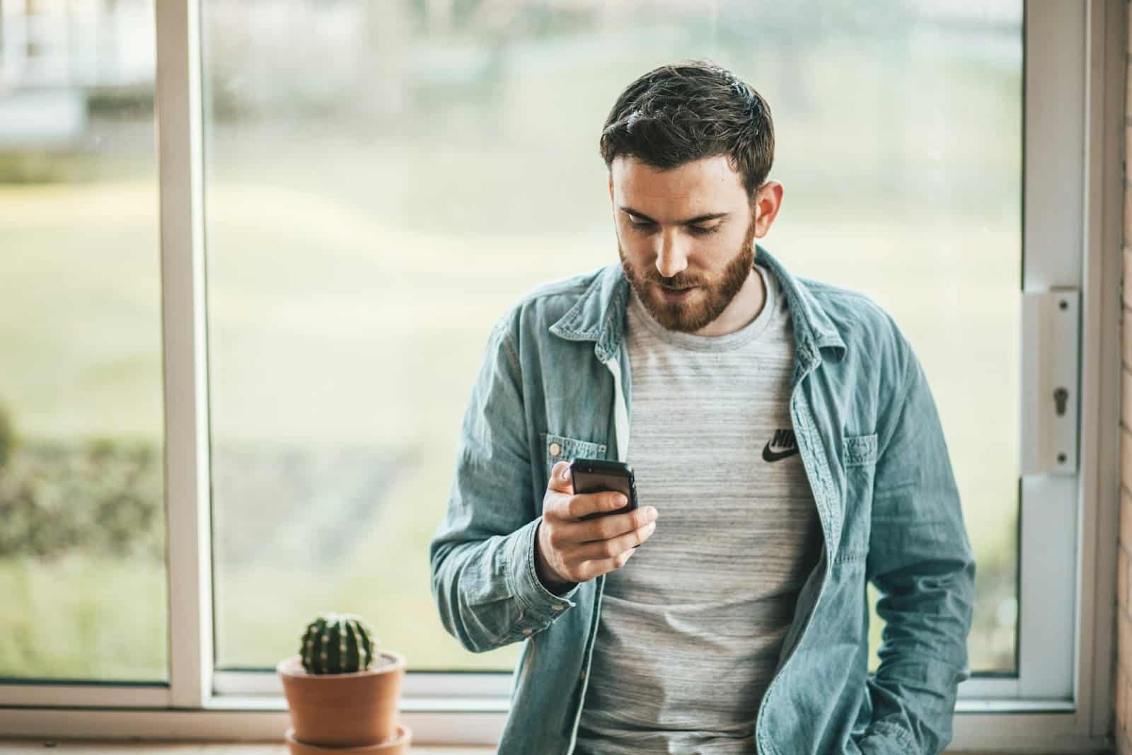 A bearded man standing while leaning against a window and looking at his phone