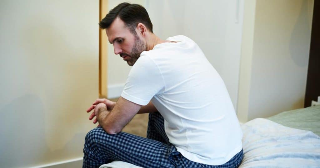 A man sitting on a bed, looking thoughtful and worried.