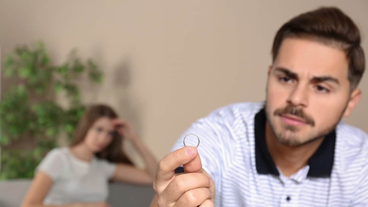A man in a striped shirt holds a silver ring while a woman sits blurred behind.