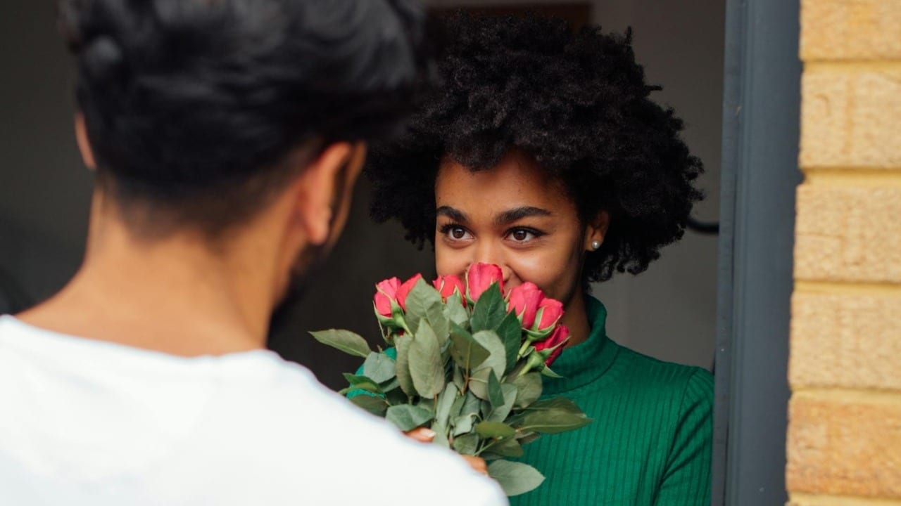 A woman in a green sweater smiles while receiving a bouquet of red roses from a man.