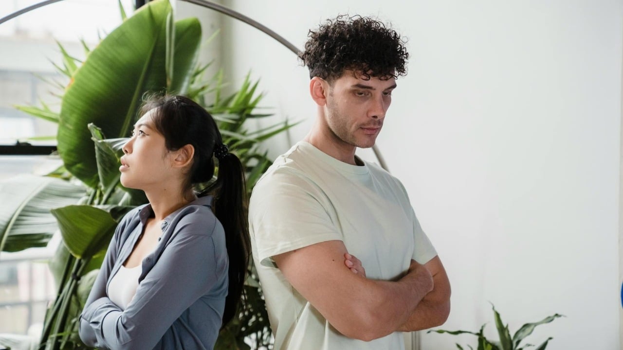 A man and woman stand back-to-back with crossed arms and somber expressions near a plant.
