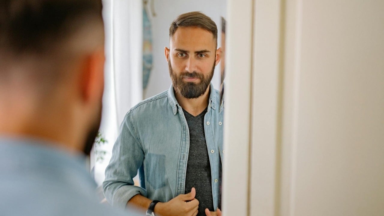 A bearded man in a denim shirt looks at his reflection in a bathroom mirror.