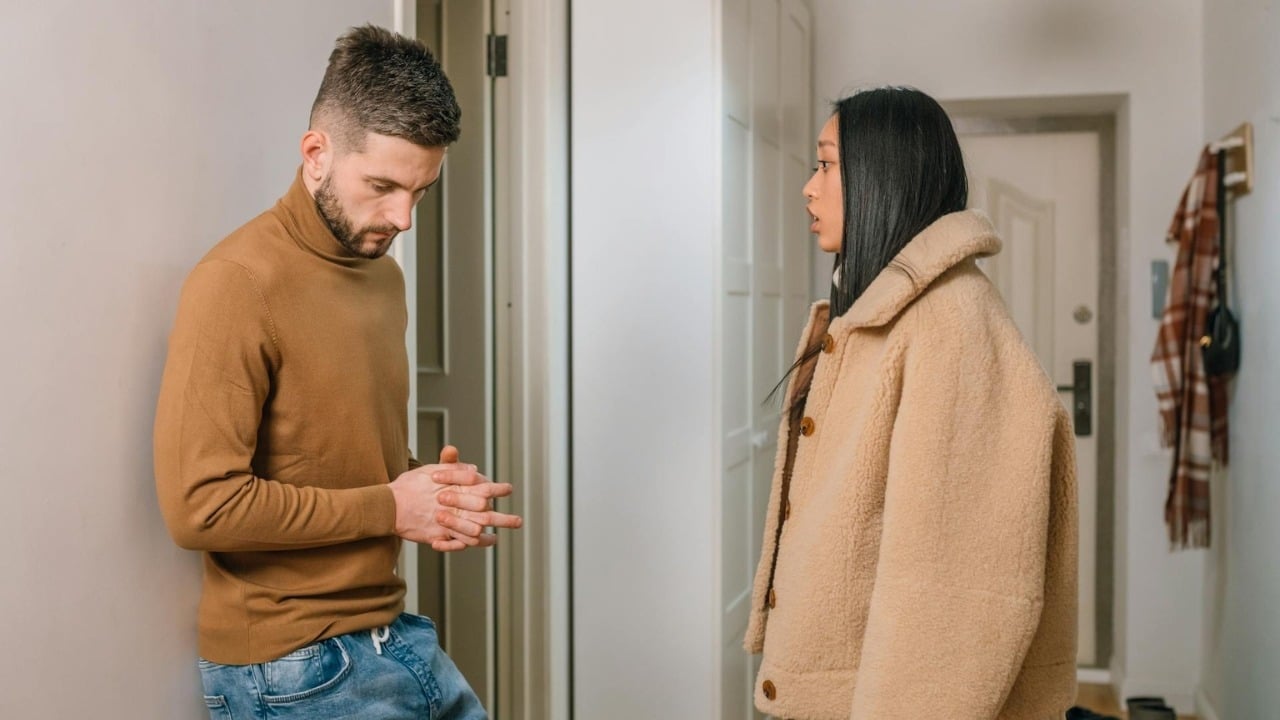 A man looks down with clasped hands while a woman in a coat speaks nearby.