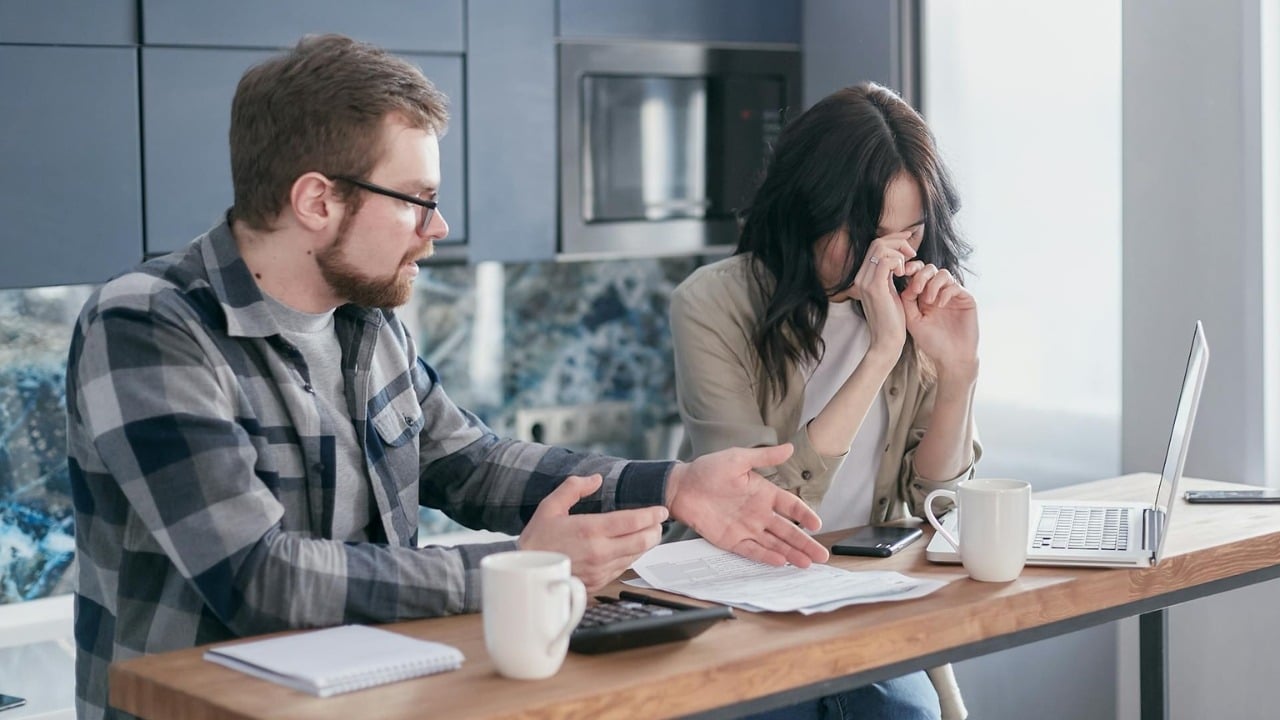 A man gestures toward paperwork while a woman beside him covers her face in distress.