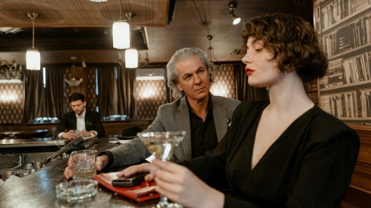 A man in a grey suit jacket looks at a woman holding a drink at a bar.