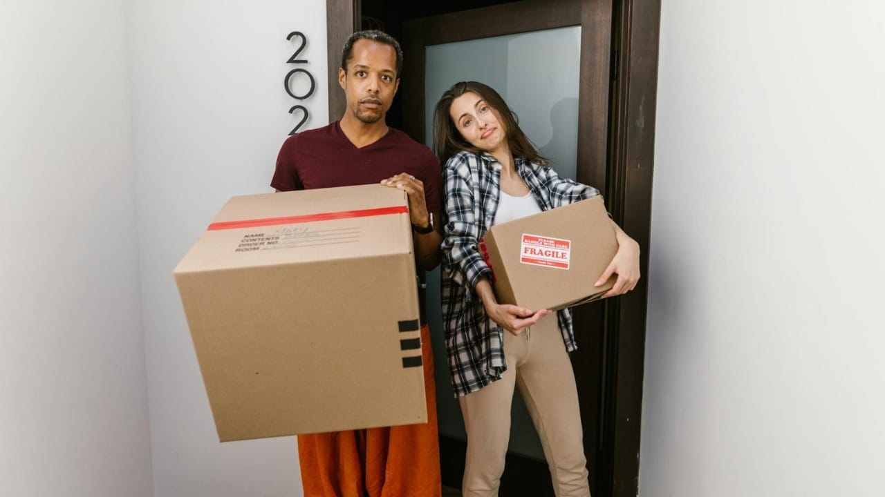 A man and woman hold cardboard moving boxes while standing in front of a doorway.