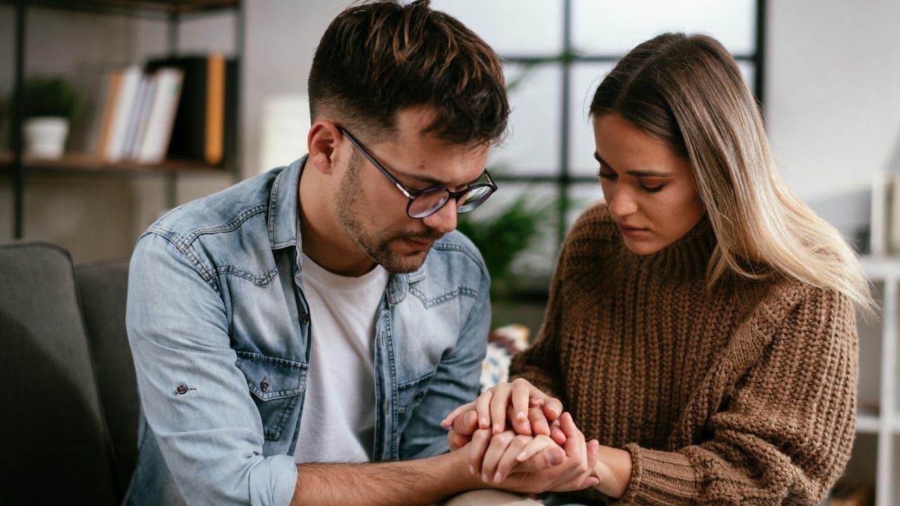 A man and woman sit close together and hold hands with somber, downcast facial expressions.