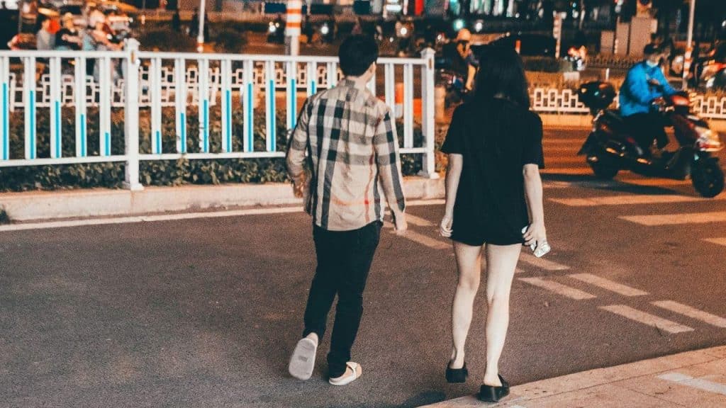 Two people walk across a street at night, viewed from behind, toward a white fence.