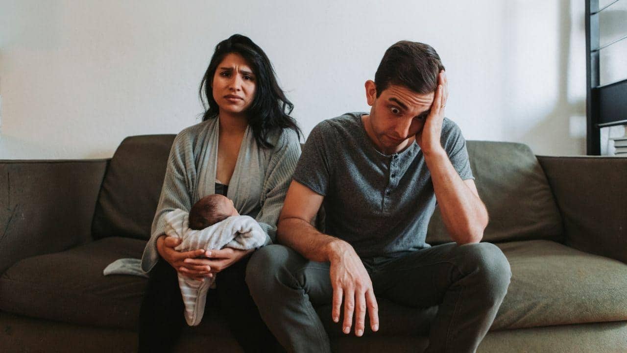 A man and a woman holding a baby sit on a couch with stressed facial expressions.