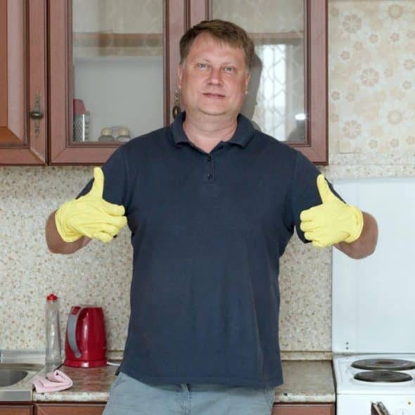 A man in a navy polo shirt and yellow gloves gives two thumbs up in a kitchen.