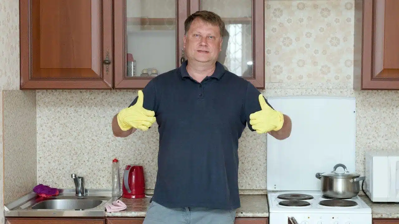 A man in a navy polo shirt and yellow gloves gives two thumbs up in a kitchen.