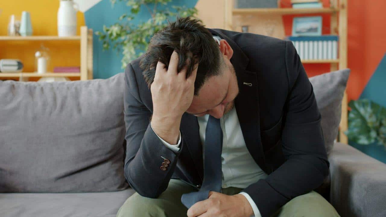 A man in a suit jacket sits on a sofa with his head in his hand.