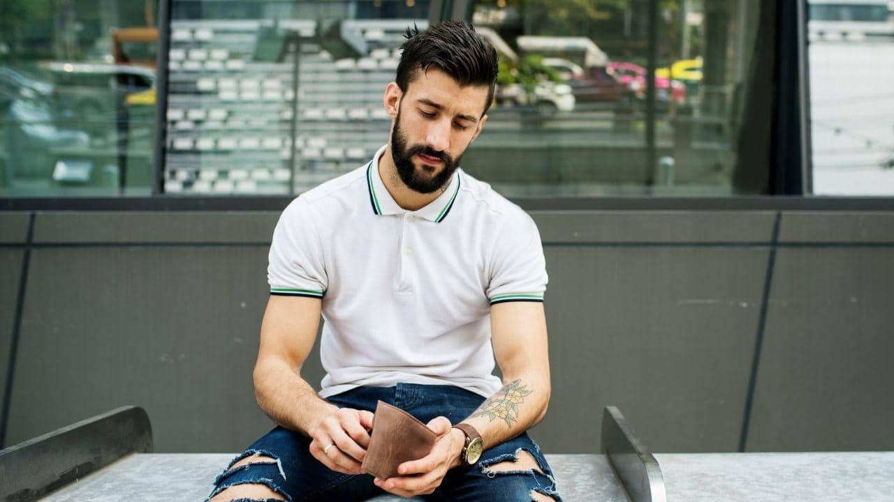 A man with a beard sits outdoors and looks down into an empty brown leather wallet.