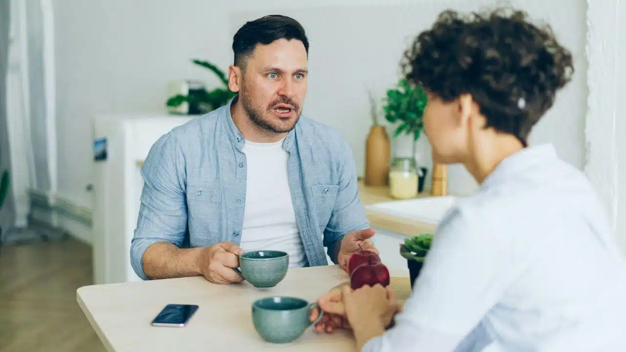 A man and woman sit at a table with mugs while the man speaks with expression.