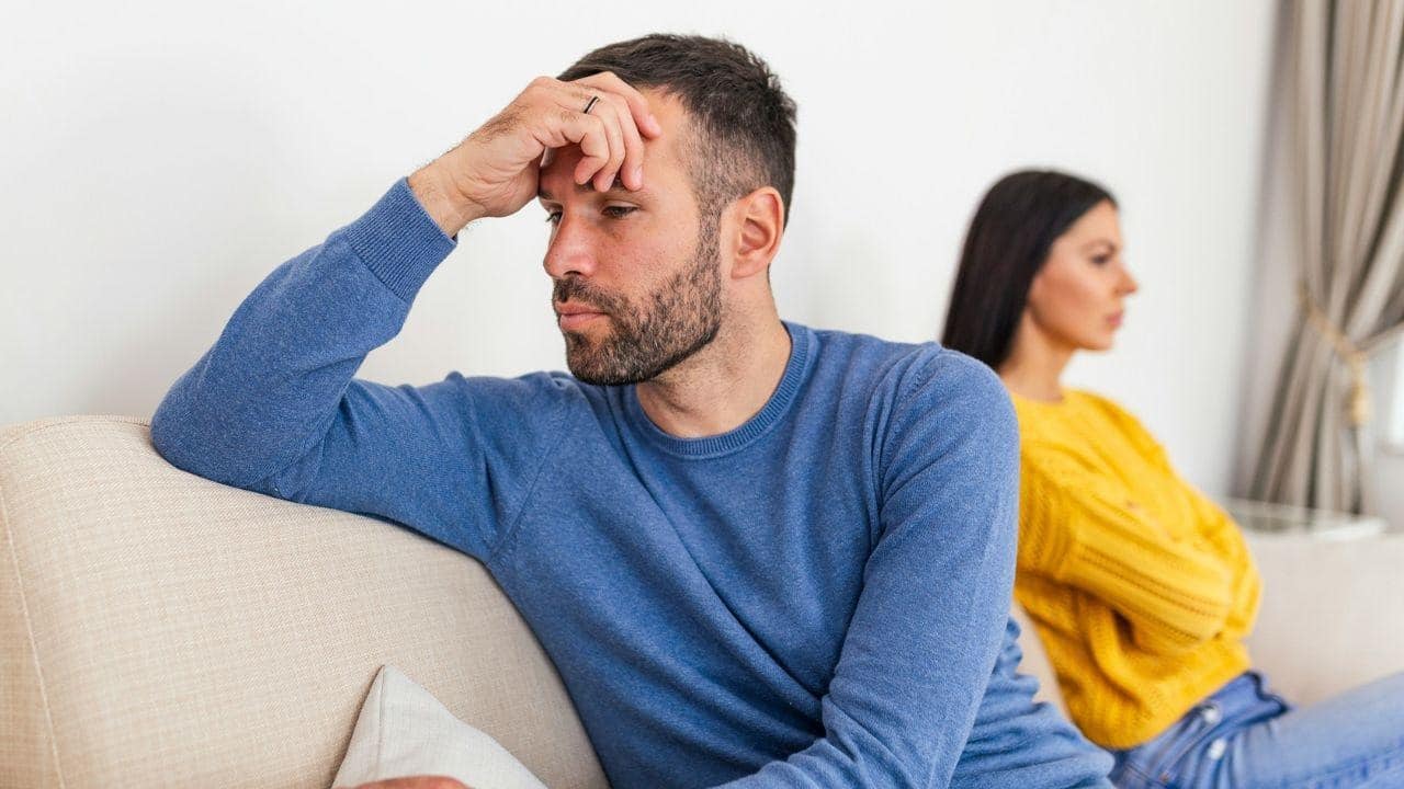 A man sits on a sofa with his hand on his head while a woman sits nearby.
