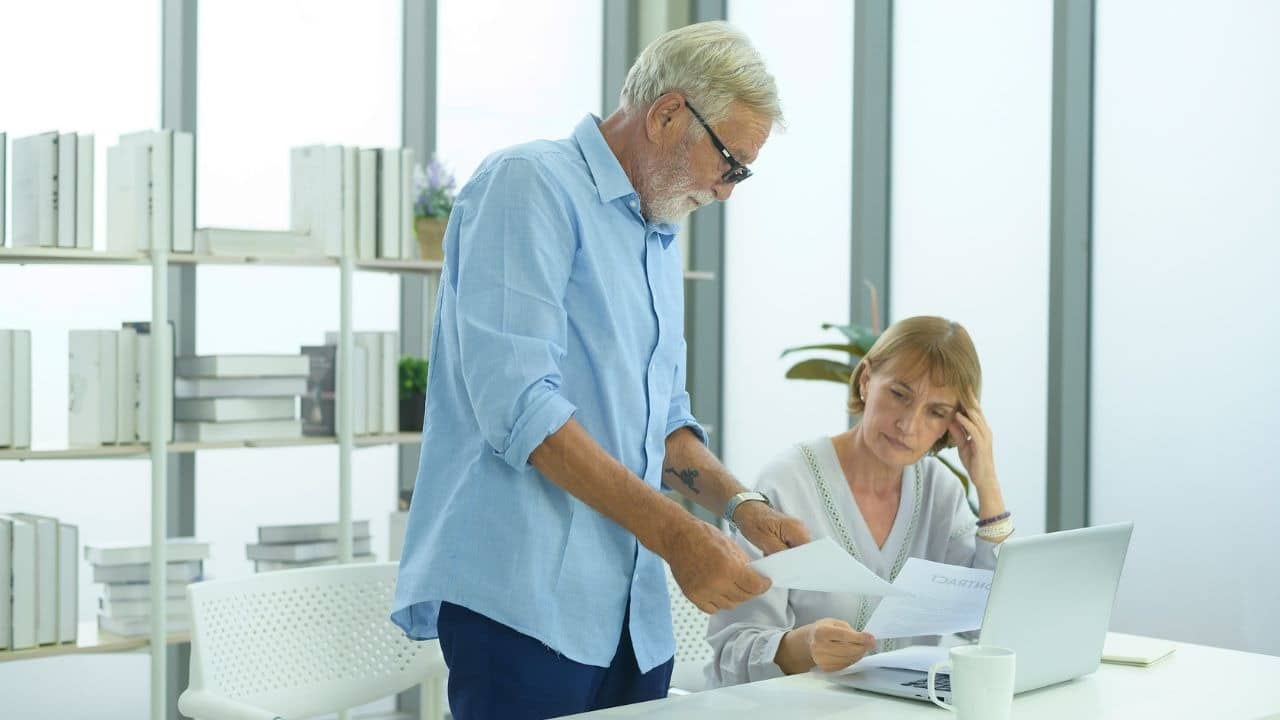 An older man stands and hands papers to a seated woman looking at a laptop computer.