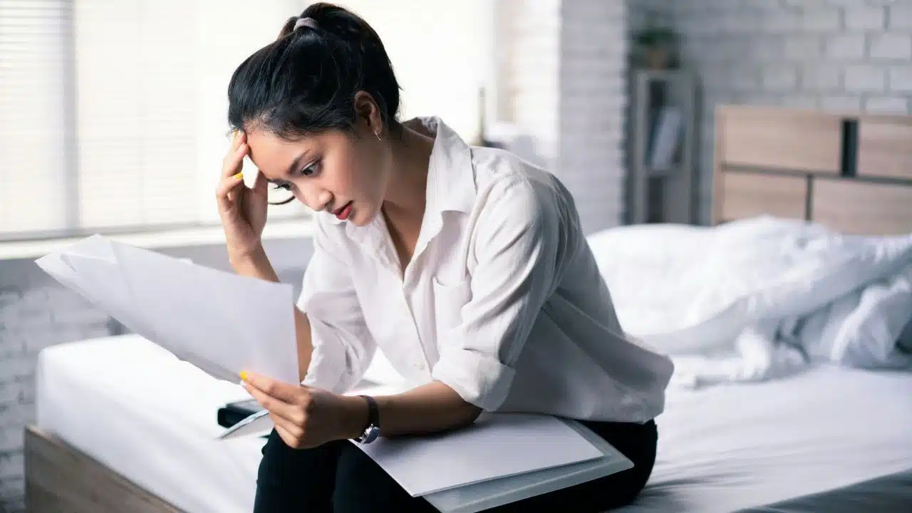 A woman sits on a bed looking at papers with her hand on her forehead.