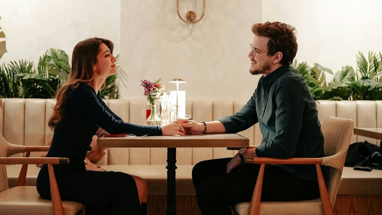 A man and woman sit at a restaurant table, holding hands and looking at each other.
