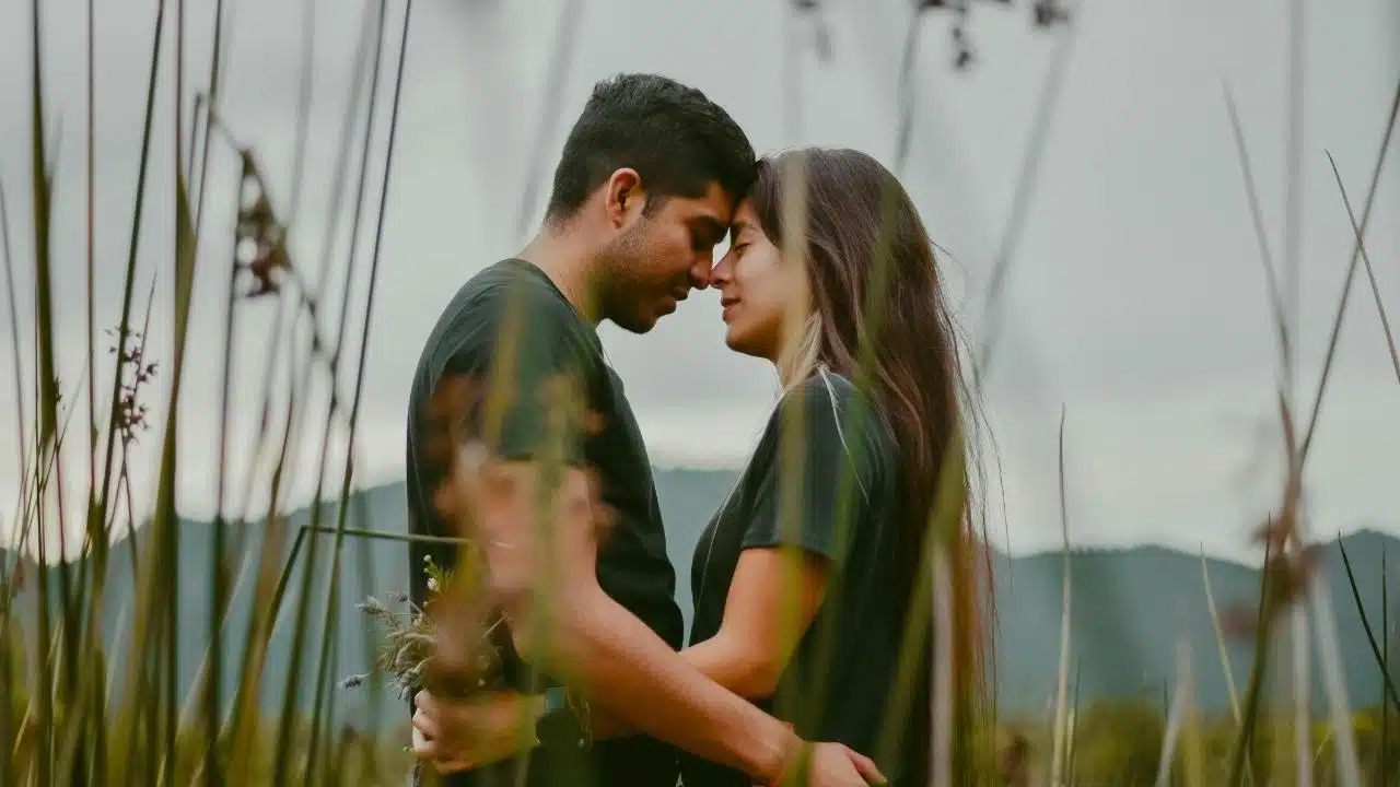 A man and woman stand in a tall grassy field with their foreheads touching together.