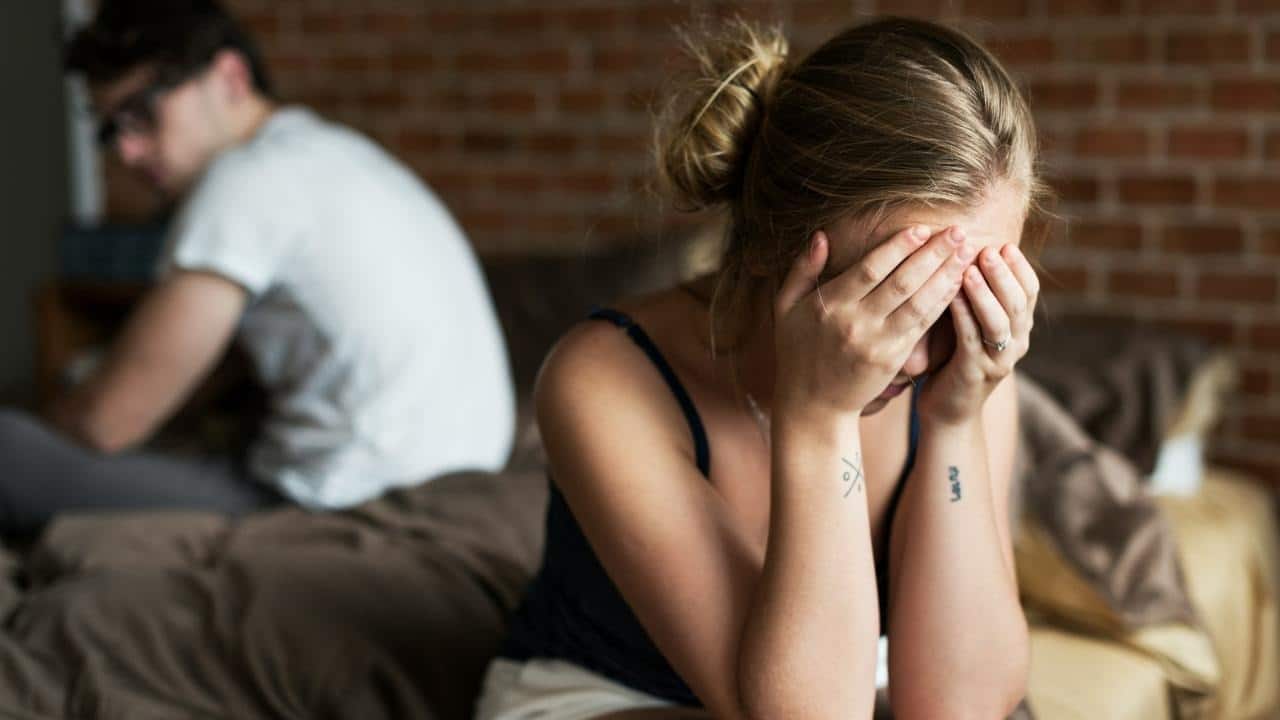 A woman sits on a bed with her head in her hands while a man sits behind.