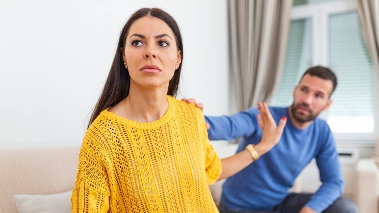 A woman in a yellow sweater looks away while holding her hand up to a man.