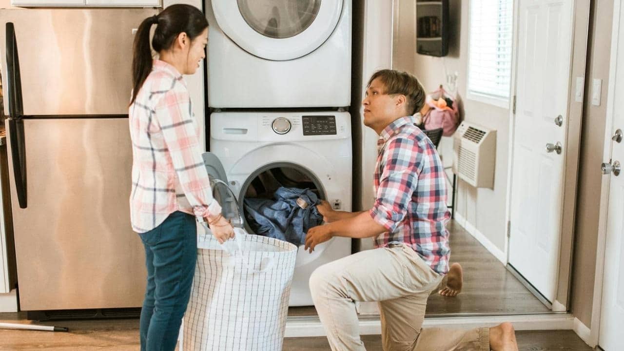 A man kneels by a washing machine while a woman nearby holds a large laundry basket.