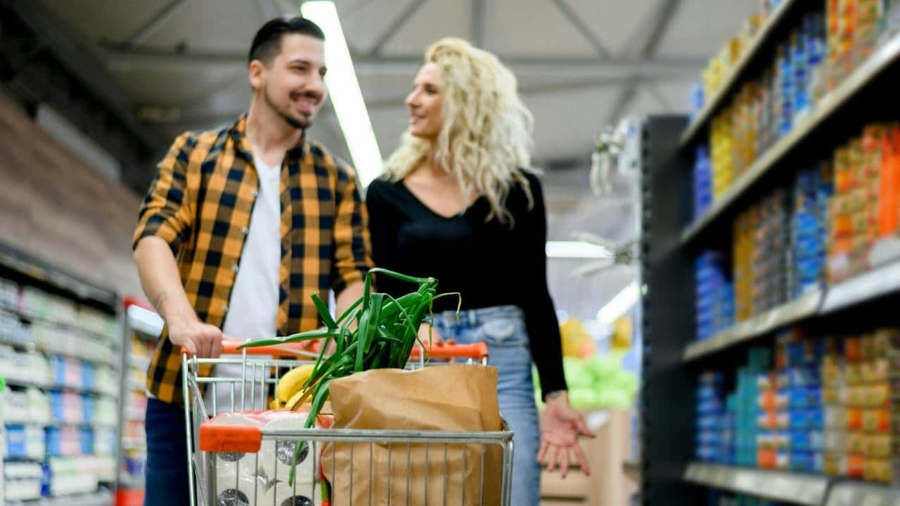 A man and woman walk through a grocery store aisle pushing a full metal shopping cart.