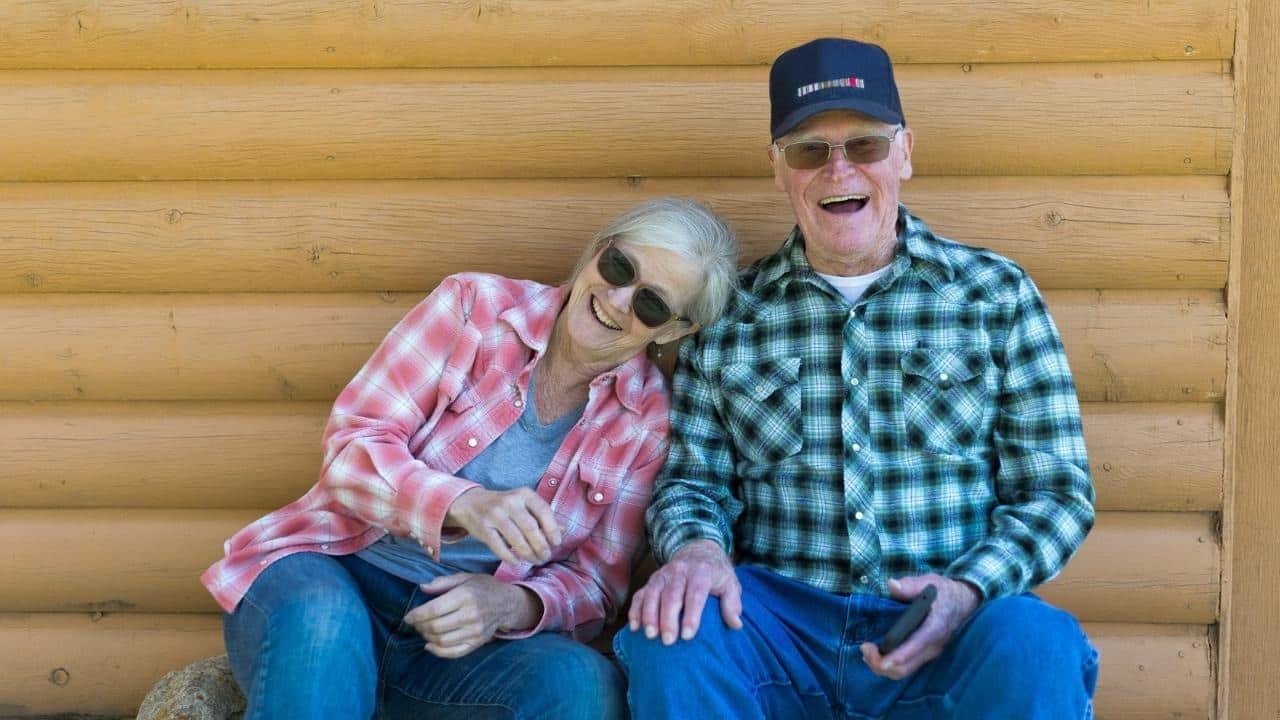 An older man and woman sit together against a log wall, laughing and smiling brightly.