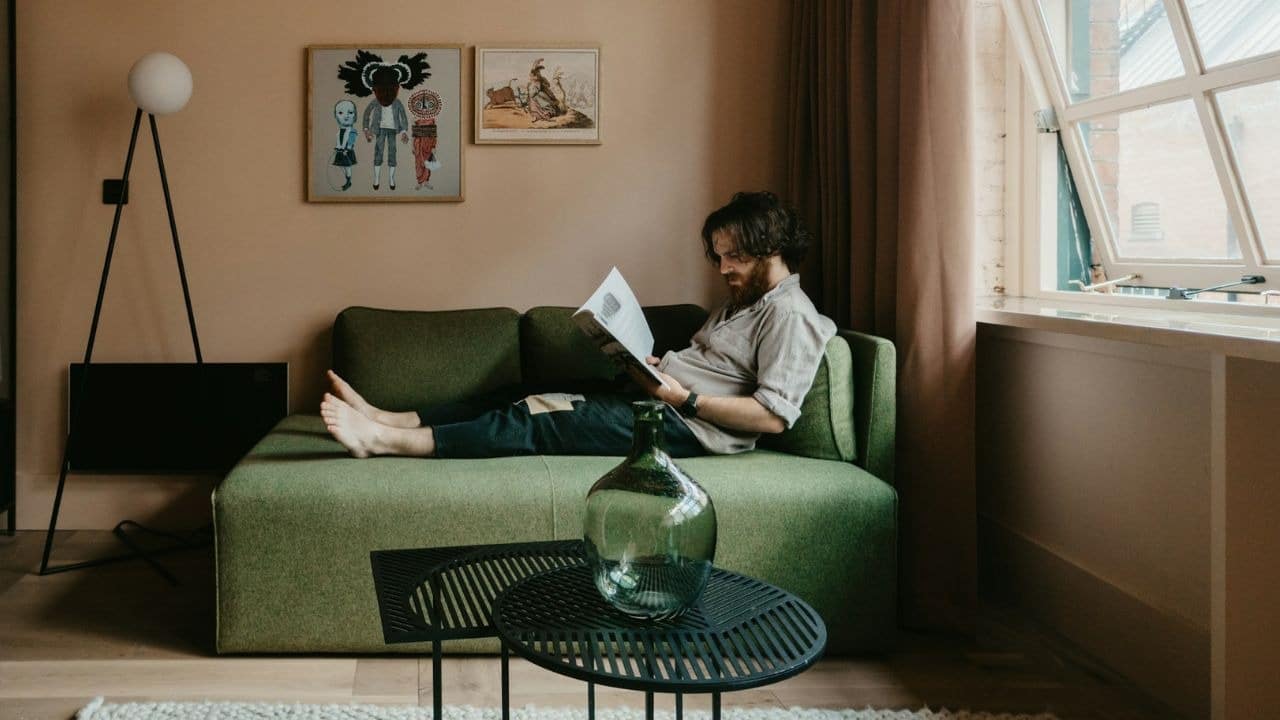 A bearded man sits alone on a green sofa indoors while reading a magazine.