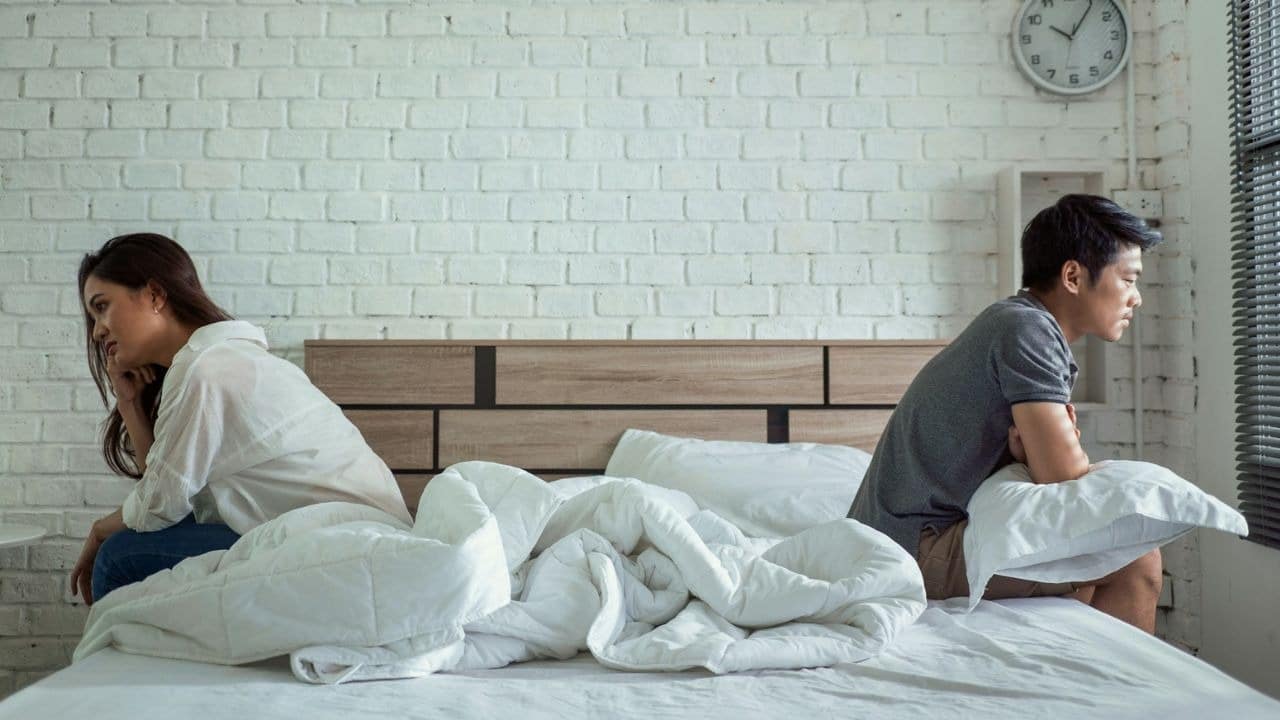 A man and woman sit on opposite ends of a bed looking in different directions.